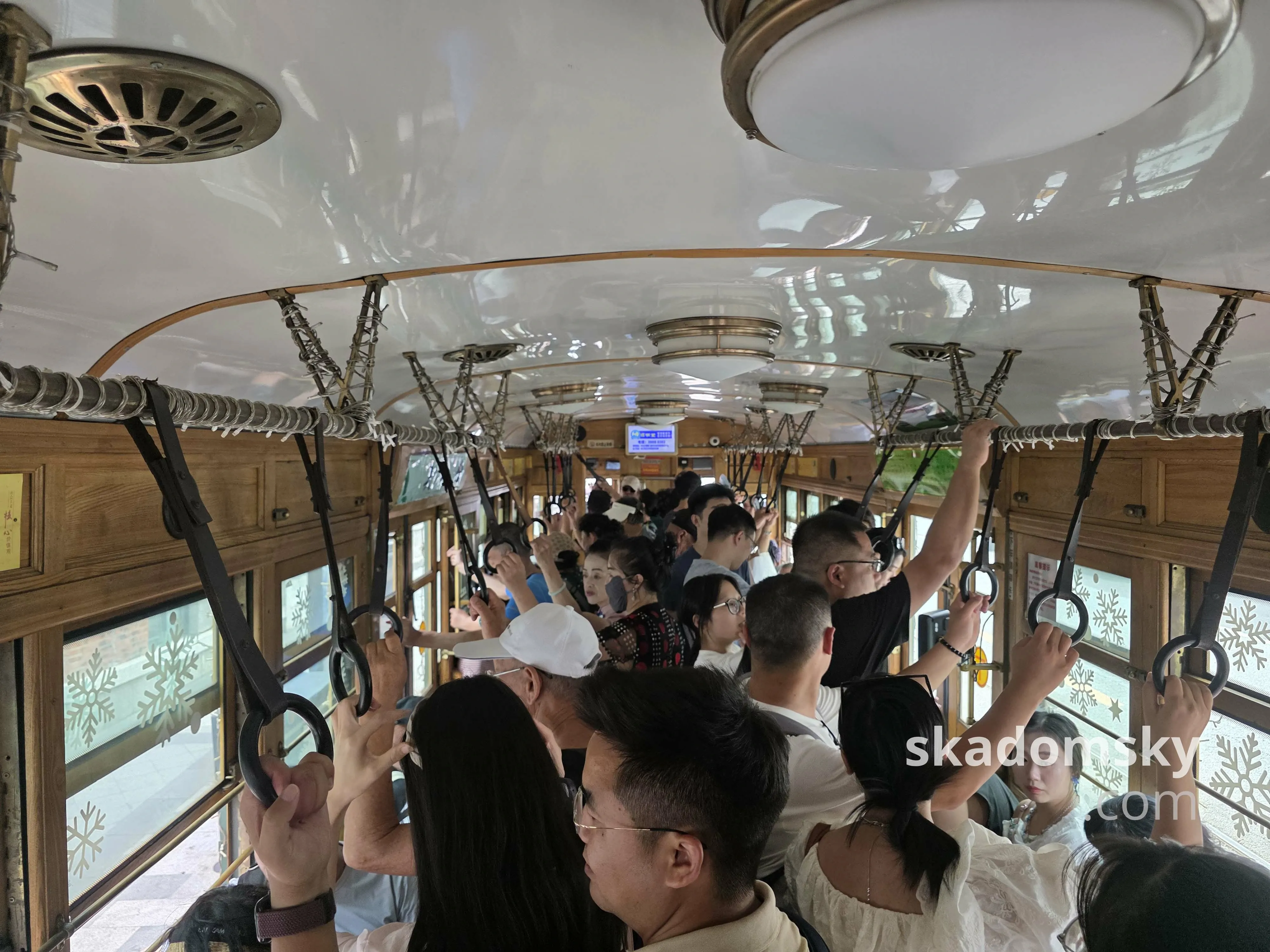 电车内部是木质结构,顶部是白色的,连着多盏白色大灯。车内熙熙攘攘,许多人握着两边的扶手。